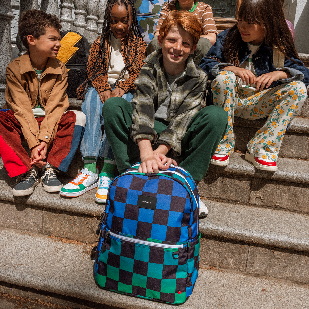 Boy sitting on stairs with friends holding STATE Bags Kane Double Pocket Blue Checkerboard